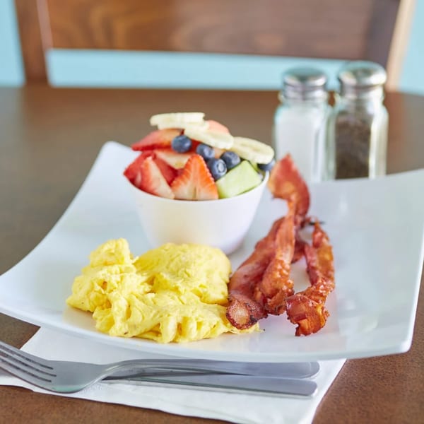 Breakfast plate with scrambled eggs, crispy bacon, and a fruit cup with strawberries, blueberries, and bananas.