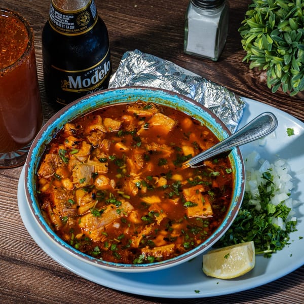 Menudo soup with cilantro, onions, lime, a Modelo beer, and a michelada on a wooden table.